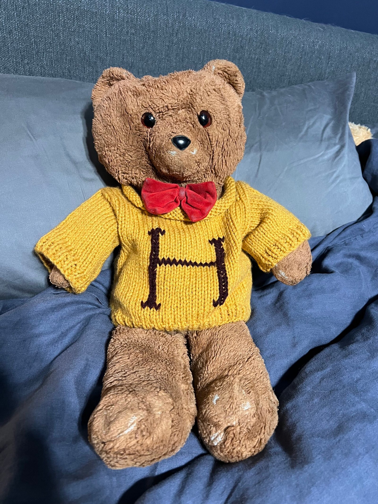A picture of the artist's own stuffed bear. A well-loved brown teddy bear with a red bow tie and yellow knitted sweater.