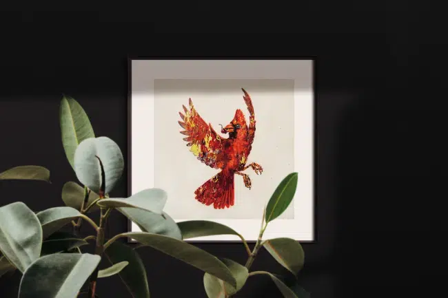 Winter Cardinal-framed mockup oin room. A drawing of a red cardinal in flight, holding a cluster of berries in its mouth. The drawing is colored over with abstract elements of paint in various colors.